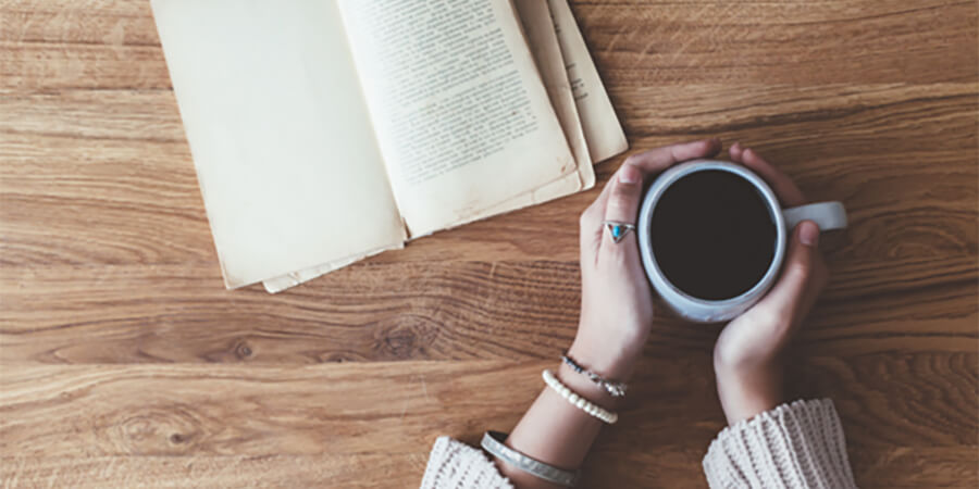 An open book and two hands wrapped around a cup of coffee.