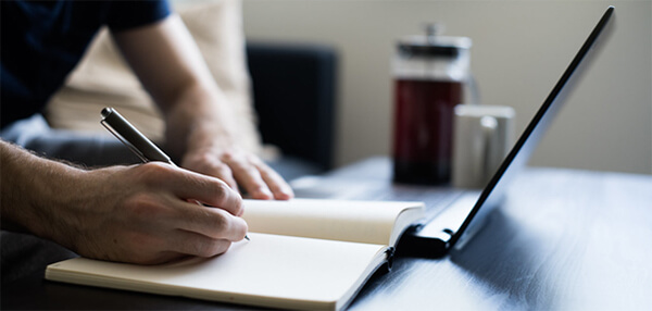 Man making notes in a notebook with a laptop and coffee.