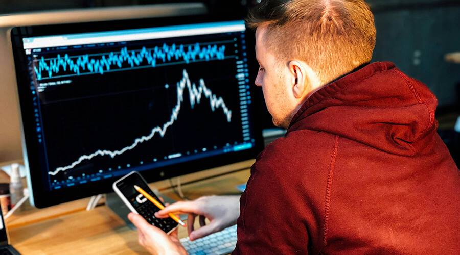 Data analyst using a calculator while looking at a graph on a monitor.