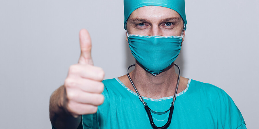 Close up of a mediacal professional wearing blue scrubs and a mask giving a thumbs up.