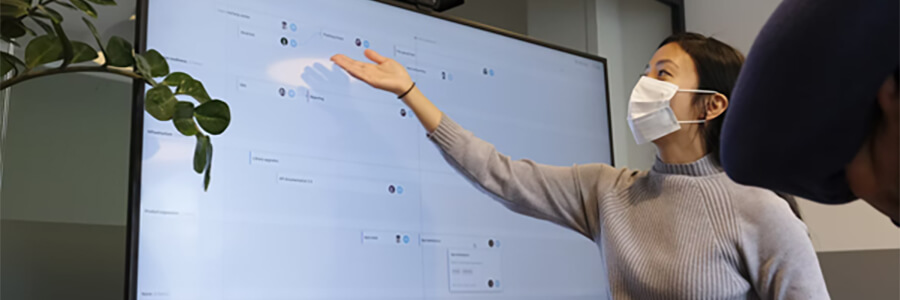 Woman wearing mask giving presentation on a large screen.