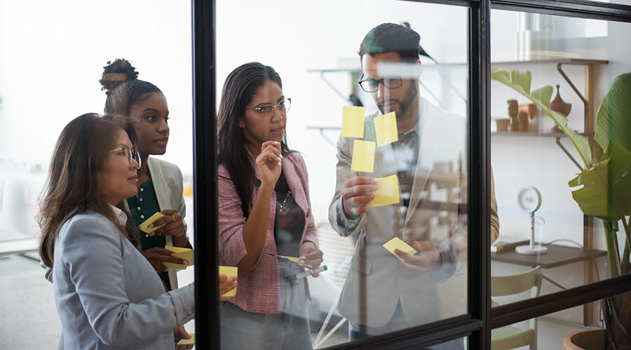 A group of people writing on Post It notes behind a glass wall.