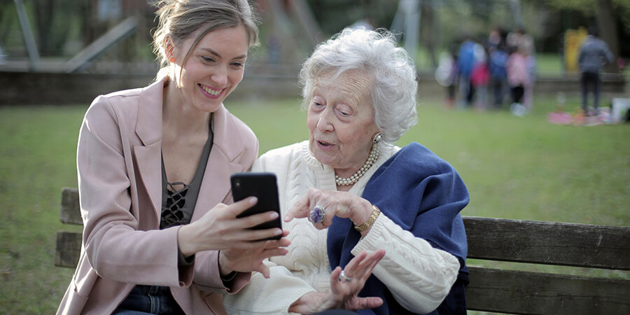 Younger woman showing an alder woman something on a smart phone while sitting on a park bench.